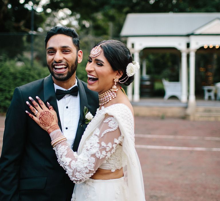 Bride & groom laugh with one another as bride wears traditional white saree and groom wears black tuxedo on their wedding day