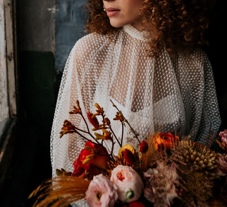 Beautiful bride with naturally curly hair and winged eyeliner holding a dried and fresh flower autumn wedding bouquet 