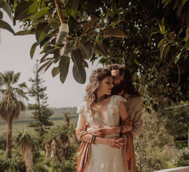 Groom wraps his arms around bride as she looks down and wears shawl around her arms