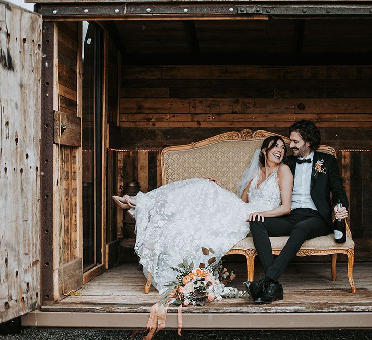 Bride in an appliqué wedding dress sitting on an ornate couch with her husband in a tuxedo at new Lake District wedding venue, Ghyll Barn 