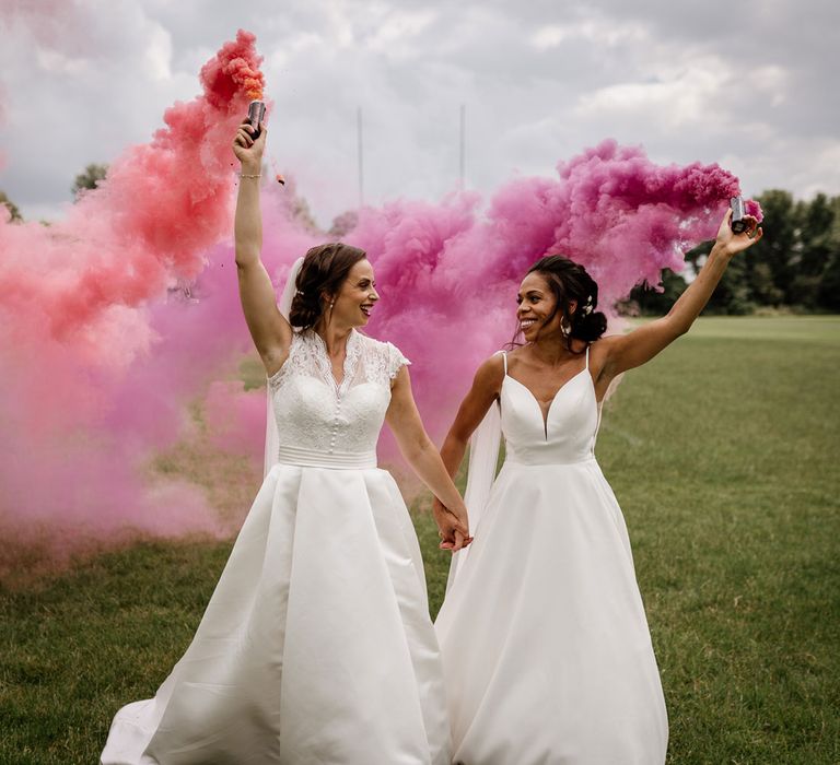 Bride in white cami wedding dress holds hands with bride in white lace top capped sleeved wedding dress on rugby pitch as they both hold up smoke bombs after wedding at The West Mill Derby