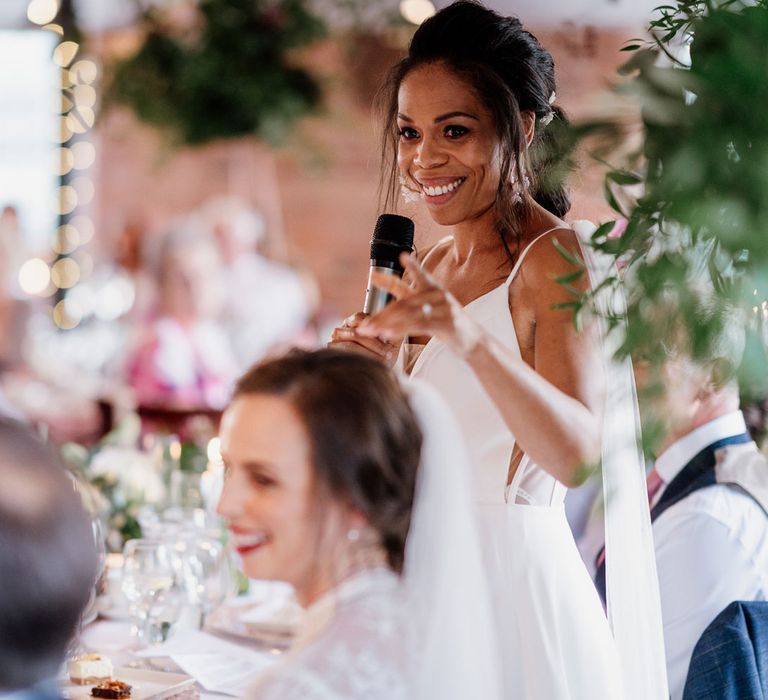 Bride in white cami wedding dress with shoulder veil details holds microphone as she makes speech during wedding reception at The West Mill Derby