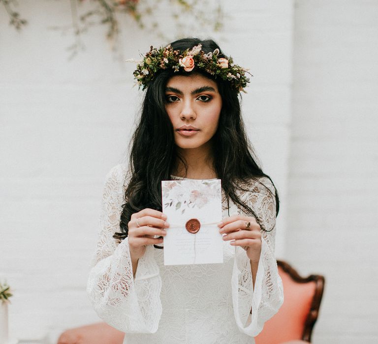 Boho bride in a lace wedding dress with long bell sleeves holding a wedding invitation with pink flower design and bronze wax seal 