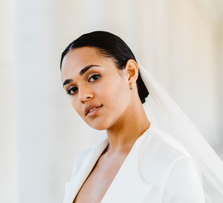 A bride wears a low cut white wedding suit and a veil in her hair, which has a central parting and is tied in a low bun.