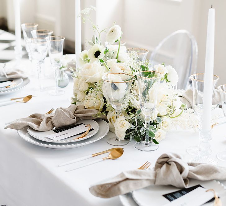 A black and white wedding tablescape. A bouquet of ranunculus, roses and anemones rests in the centre of the table. The place settings are double plats, gold cutlery and gold rimmed glassware. Tied napkins rest on the white plates. 