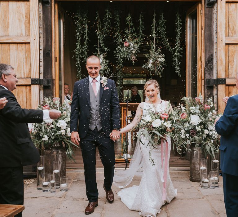 Groom in dark three piece suit with grey waistcoat and pink tie holding hands with bride in lace Justin Alexander wedding dress holding pink and white bridal bouquet walk back down the aisle whilst guests throw confetti at Tythe Barn wedding 