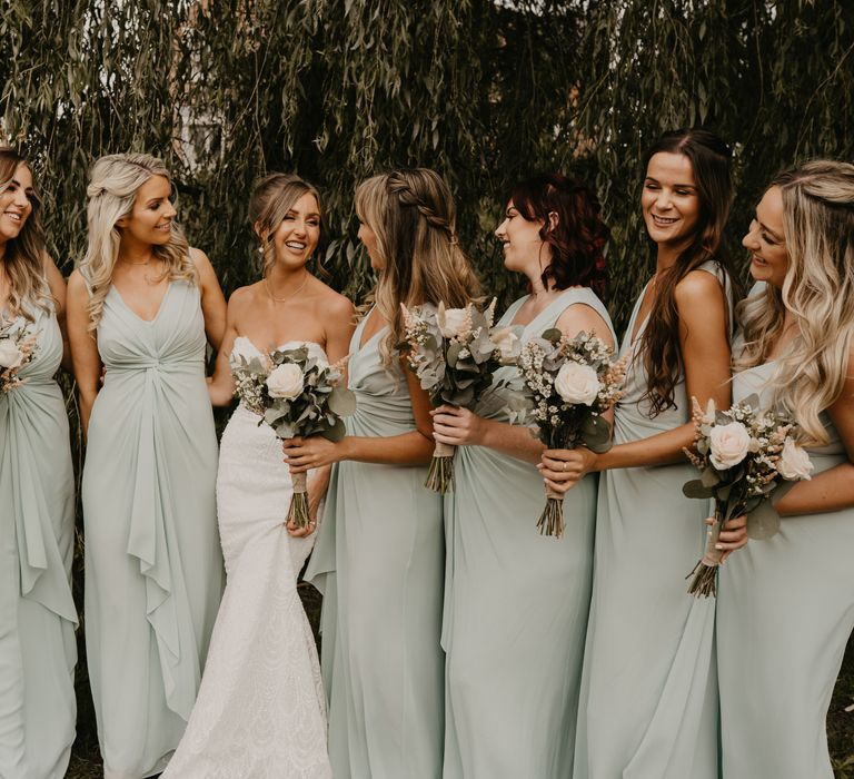 Bride stands with her bridesmaids who wear sage green dresses and carry white floral bouquets | Mark Bamforth Photography