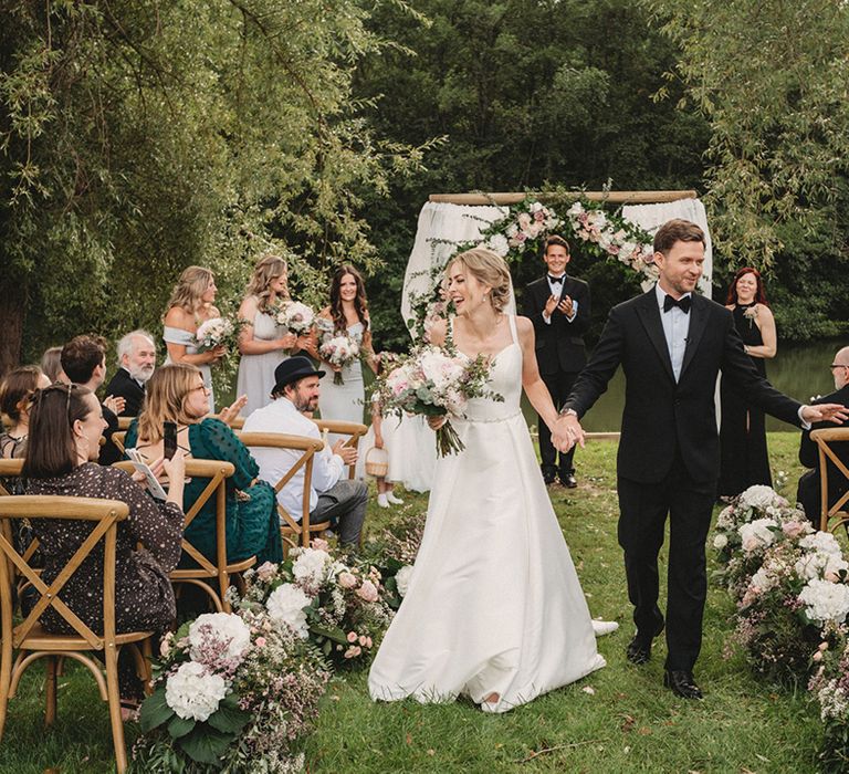 Bride in a Stella York wedding dress and groom in a tuxedo walking up the pink and white flower lined aisle at Chateau Lagorce 