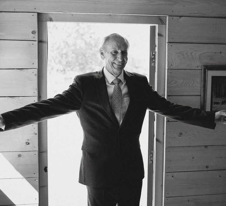 Father of the bride in suit and tie holds arms open wide and glass of sparkling wine at doorway before tipi wedding at Inkersall Grange Farm