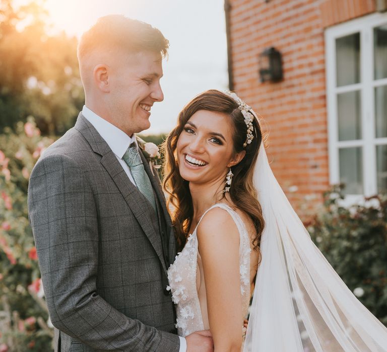 Groom in grey checked three piece suit and mint green paisley tie smiles down at bride in applique Berta wedding dress, headband and veil as they stand in garden for summer wedding at Primrose Hill Farm