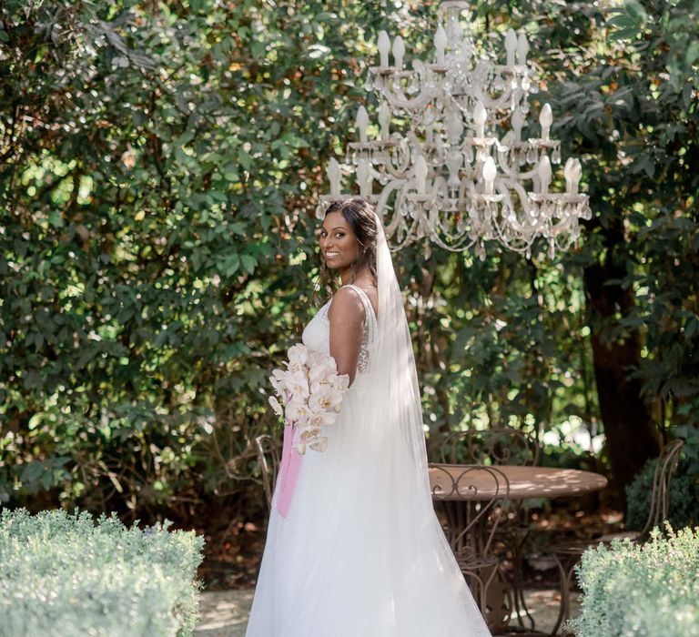 Indian bride stands outdoors in Lake Garda in front of chandelier and holding her bridal bouquet