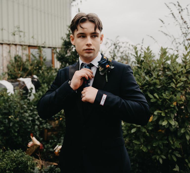 Groom in dark blue suit, floral tie and floral buttonhole outside for rustic farm wedding