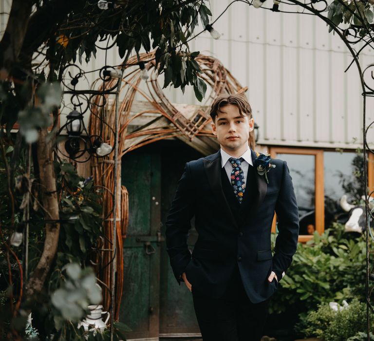 Groom in blue suit, floral tie and rose buttonhole walks through garden for rustic farm wedding in Lancashire