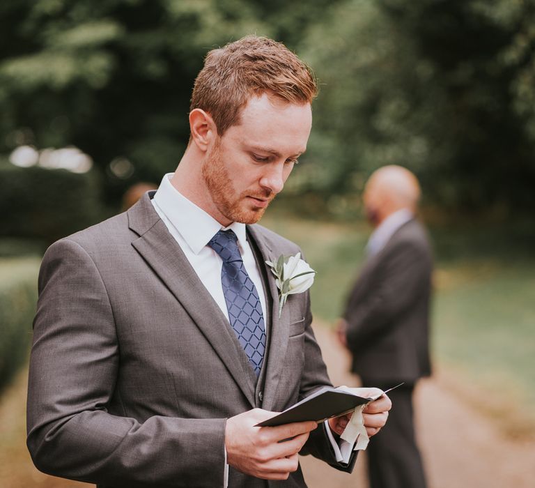 Groom in grey three piece suit, blue tie and whit rose buttonhole reads vows before outdoor wedding ceremony