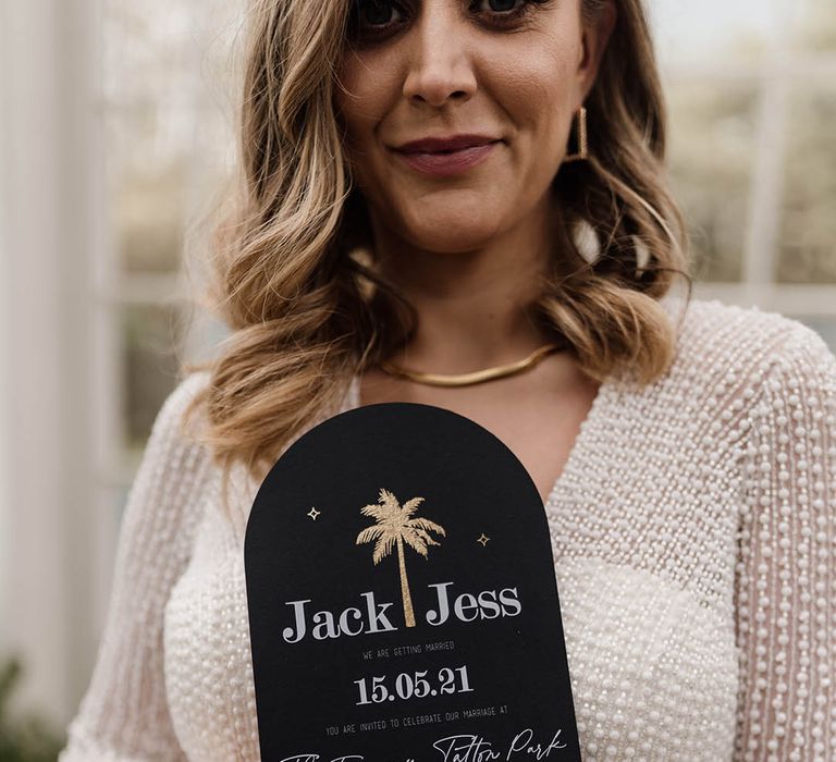 Bride in a beaded wedding dress with pink wedding nails holding a black wedding invitation with white contemporary font and gold foil palm tree design