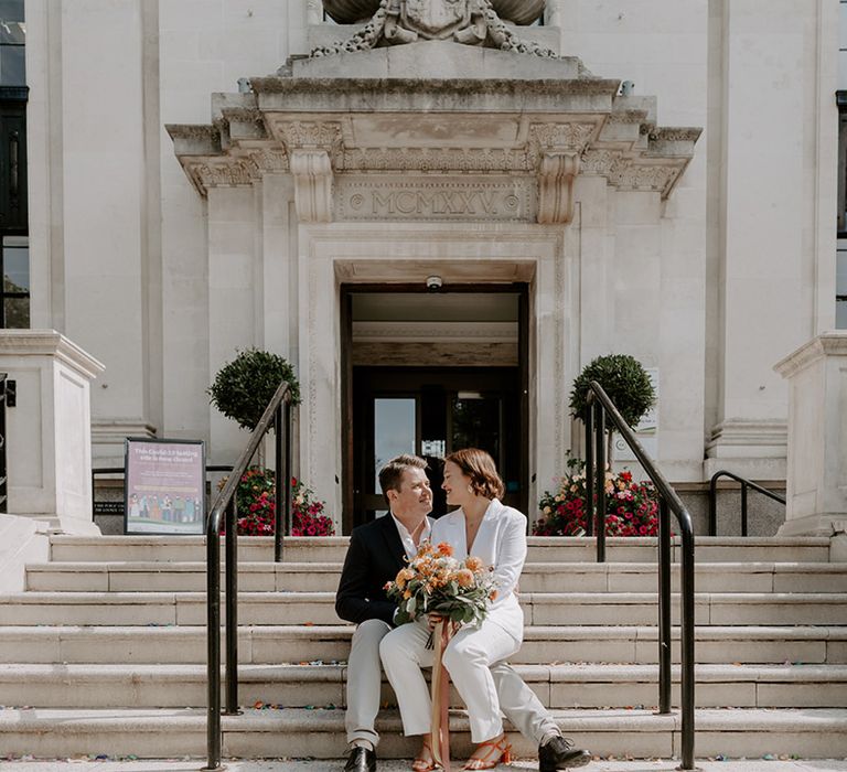 Bride and groom sitting on the steps at Islington Town Hall with bride holding an orange flower bouquet