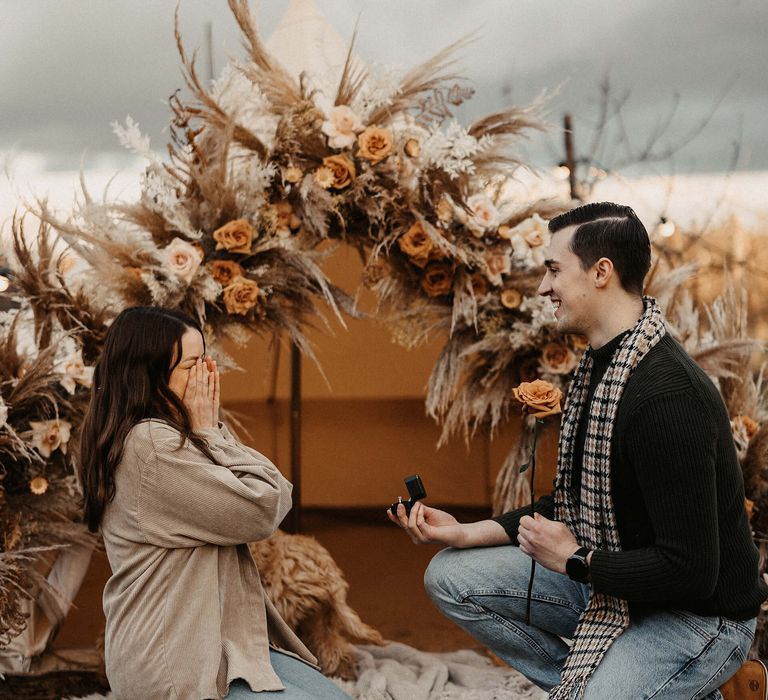 Groom-to-be down on one knee proposing to his girlfriend at their bell tent, campfire marriage proposal 