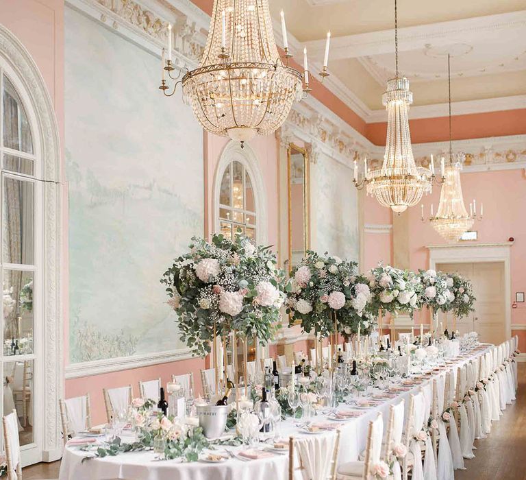 Danesfield House reception complete with grand chandeliers, long table lined with white cloth, large floral bouquets filled with blush pink and white flowers