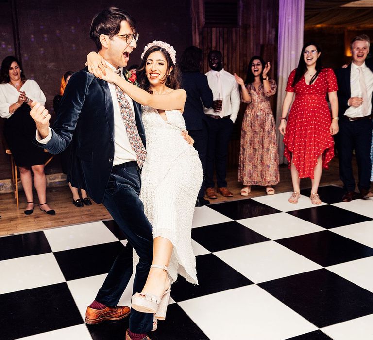 Bride in beaded white midi Dres and headband dances with groom in dark blue suit on black and white dance floor during evening wedding reception