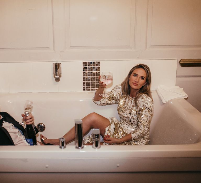 Bride & groom sit in bathtub on their wedding day and hold champagne glasses 