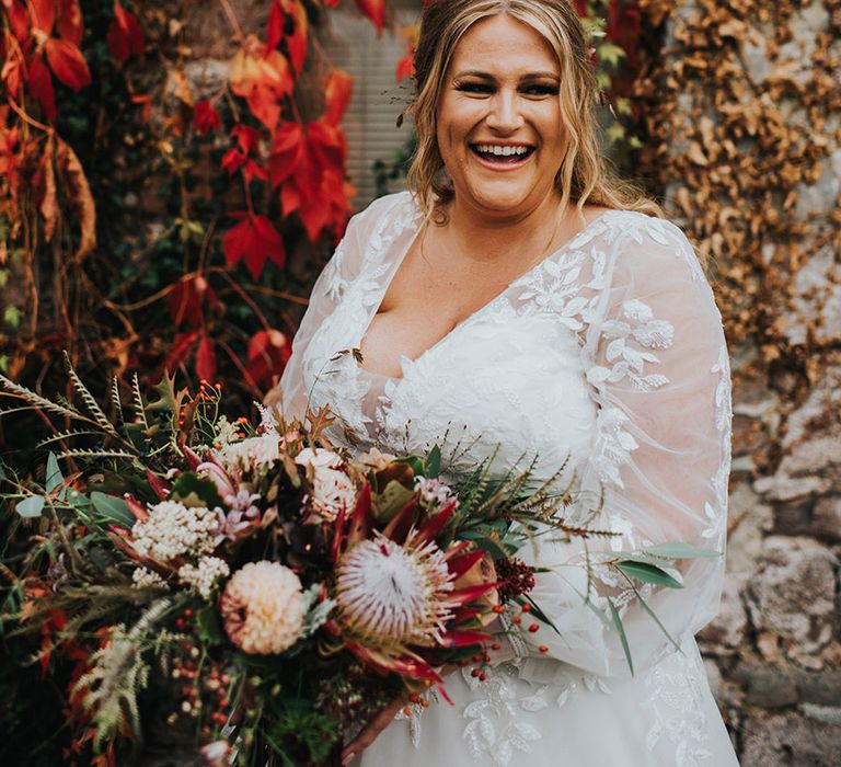 Autumn wedding with curvy bride in a V neck wedding dress with long sleeves holding her king protea bouquet 