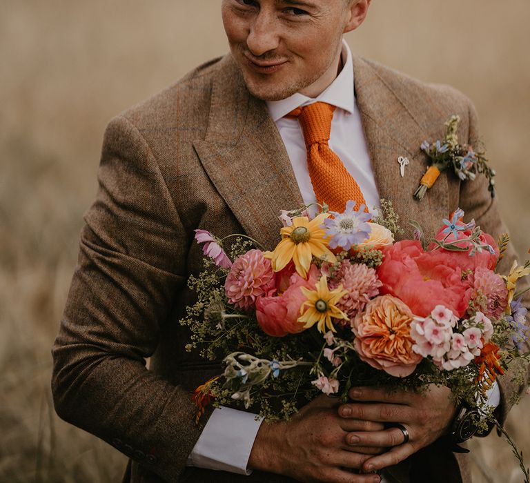 Groom in brown suit and orange tie makes a cheeky face as he holds the bridal bouquet 