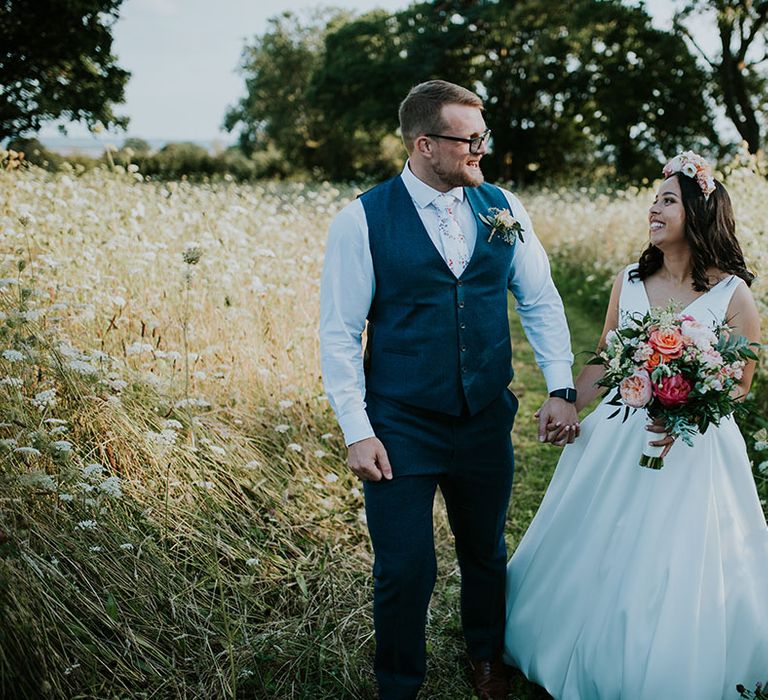 Smiling bride and groom have their fingers intertwined as they walk in the pretty grounds around St Tewdrics House 