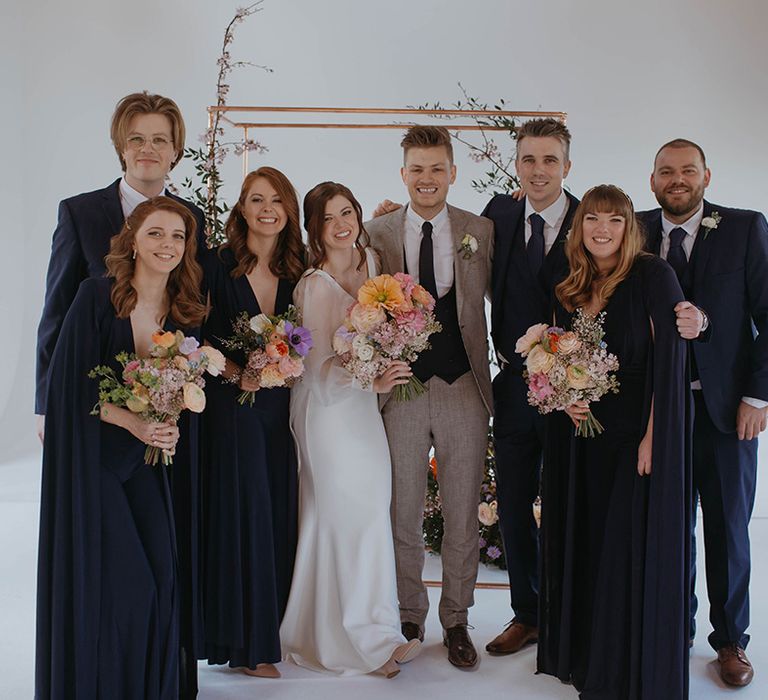 Wedding party earring navy blue smiles with the bride in wedding dress with sheer sleeves and groom in Reiss suit in front of copper frame altar