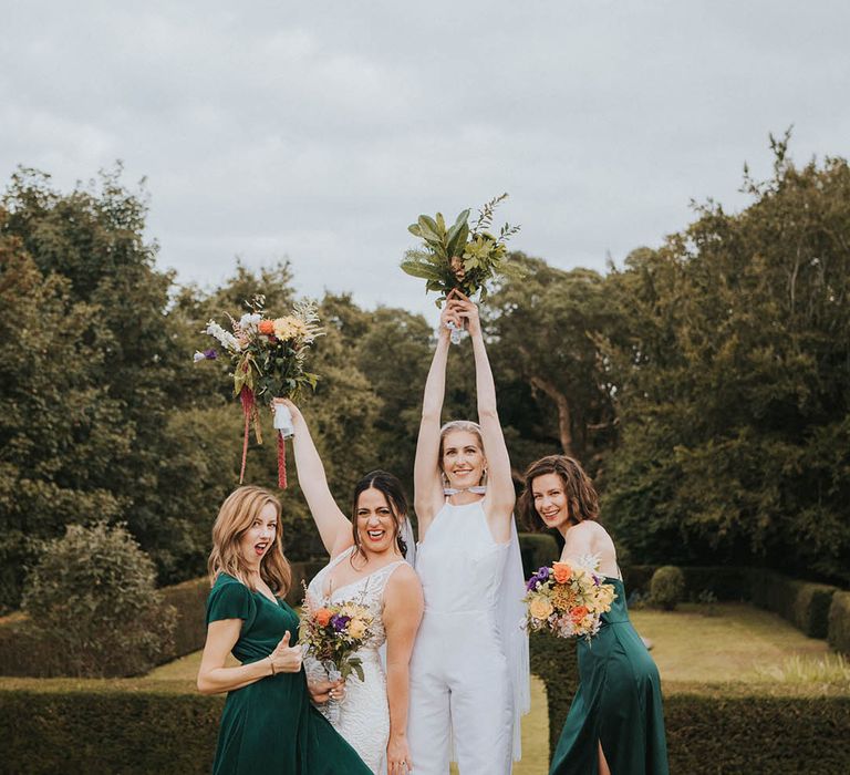 Bridesmaids in different style forest green dresses and colourful bouquets with brides in a jumpsuit and cape and lace wedding dress