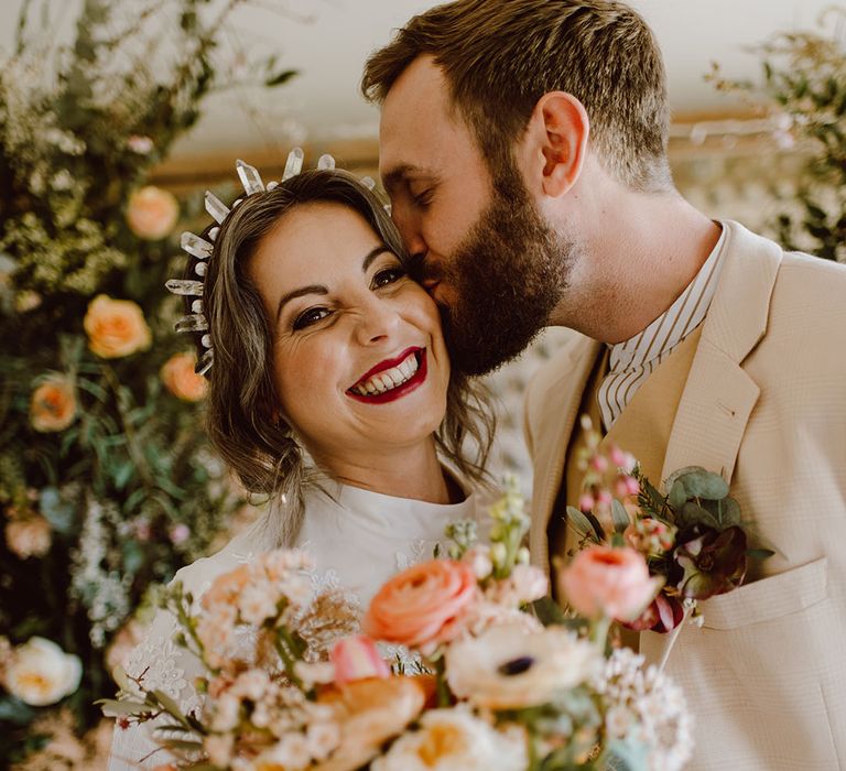 Groom in a beige suit kissing his bride in a crystal headband wearing red lipstick 