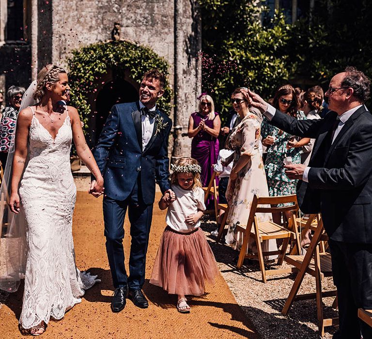 Bride in lace wedding dress and groom in blue tuxedo with their daughter in a pink tulle skirt and white flower crown have confetti exit from their wedding 