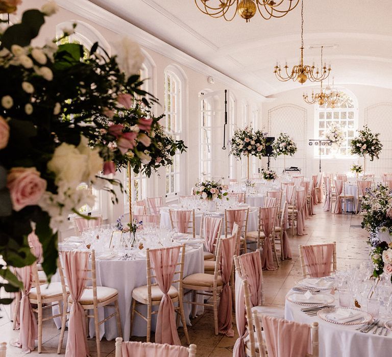 White reception room at Goldney Hall with chair backs decorated with pink chiffon material and tables decorated with tall pink rose centrepieces and gold chandeliers 