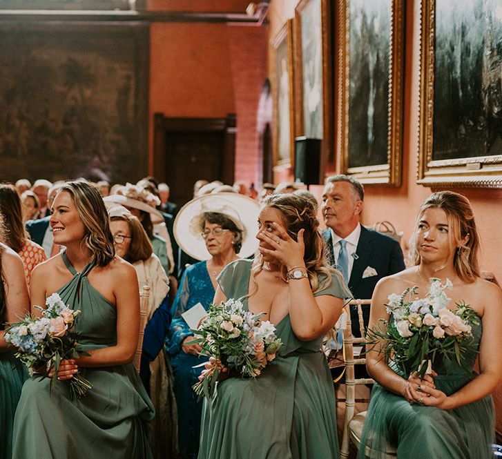 Bridesmaids wearing green bridesmaid dresses in different styles and holding pastel bouquets become emotional during wedding ceremony