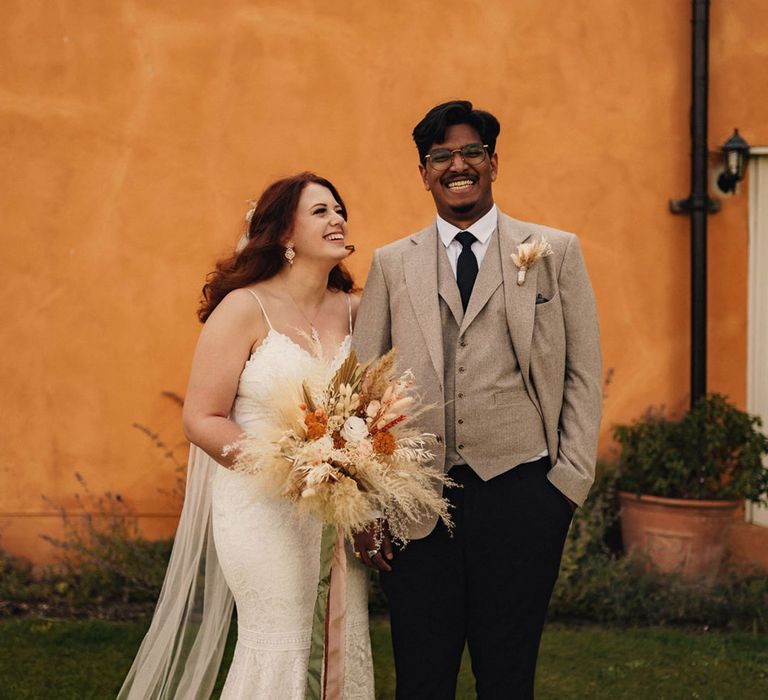 Bride in lace wedding dress with groom in grey suit with bride carrying dried grass flower bouquet 