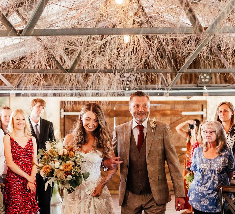 Father of the bride in brown checkered suit with a red tie walks the bride in a boho off the shoulder wedding dress down the aisle 