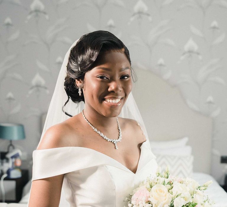 Black bride wears fitted off-the-shoulder wedding dress complete with floor-length veil and matching diamond jewellery 