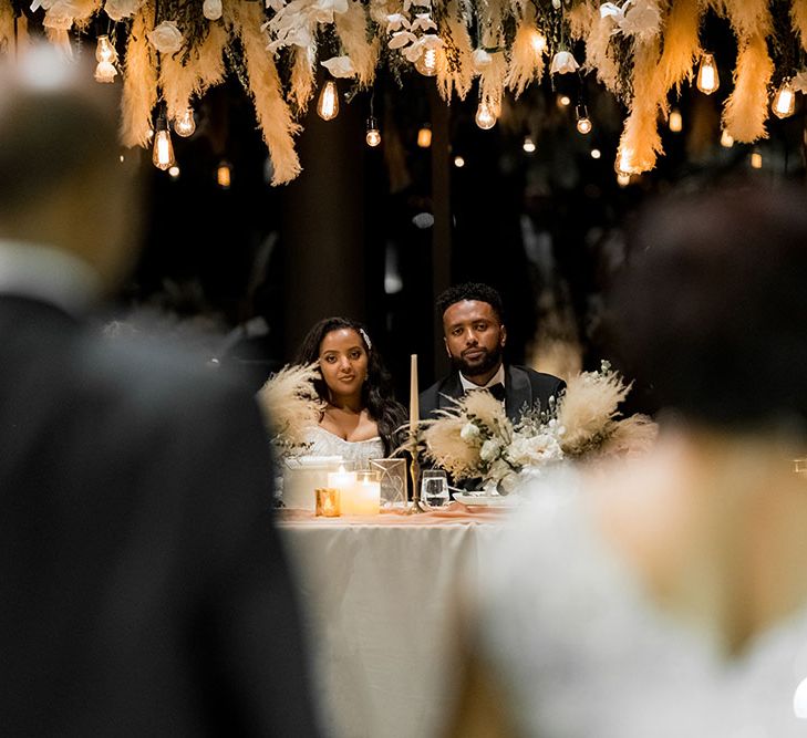 Festoon lighting hangs from pampas grass as bride & groom listen during wedding speeches 