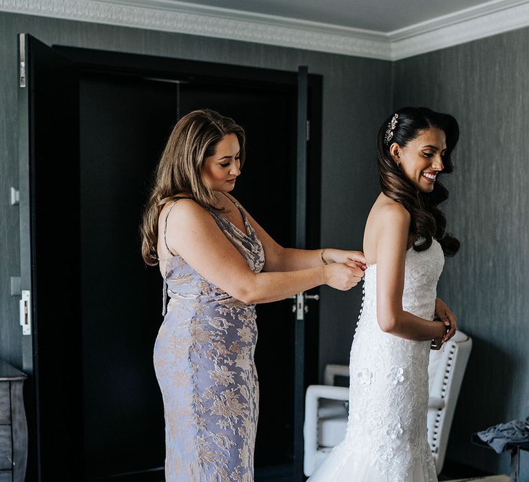 Bridesmaid wears lavender bridesmaid dress and does up buttons to the back of brides lace wedding dress 