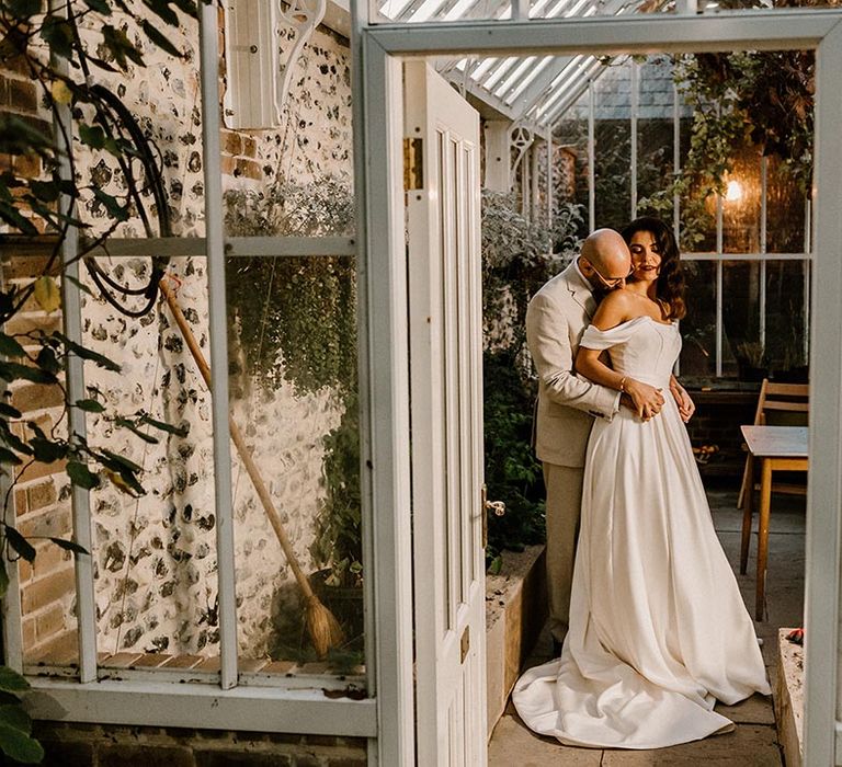 Groom embraces the bride from behind as they stand in the glasshouse at the Chapel House Estate 