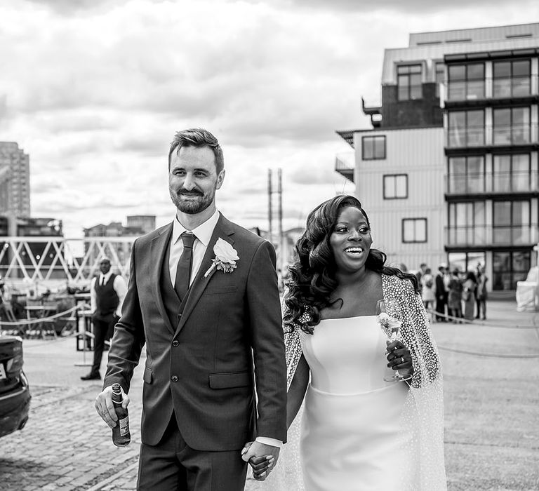 Groom wearing three-piece suit and white floral buttonhole walks alongside his bride in tulle wedding cape 