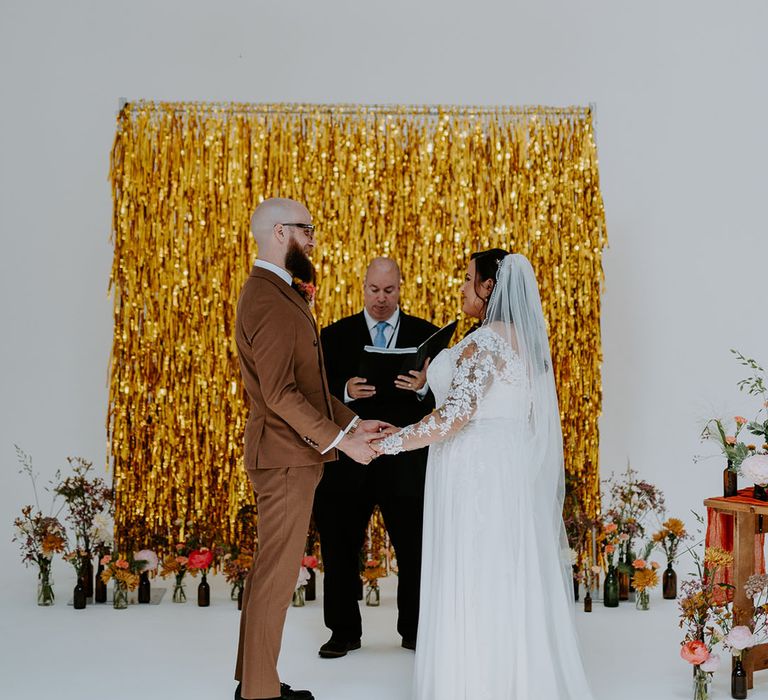 Bride & groom stand in front of gold streamers wedding decor during ceremony 