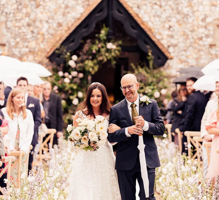 Father of the bride in a black and white suit with a satin brown tie walks the bride in an applique wedding dress down the aisle 