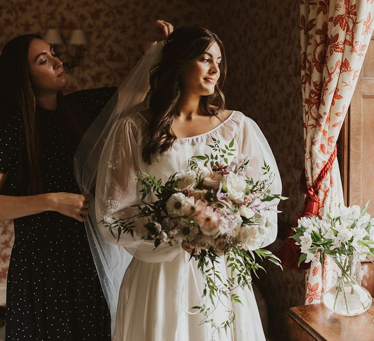 Bride with curled brown hair gets her veil put in holding a pink and white wedding bouquet 