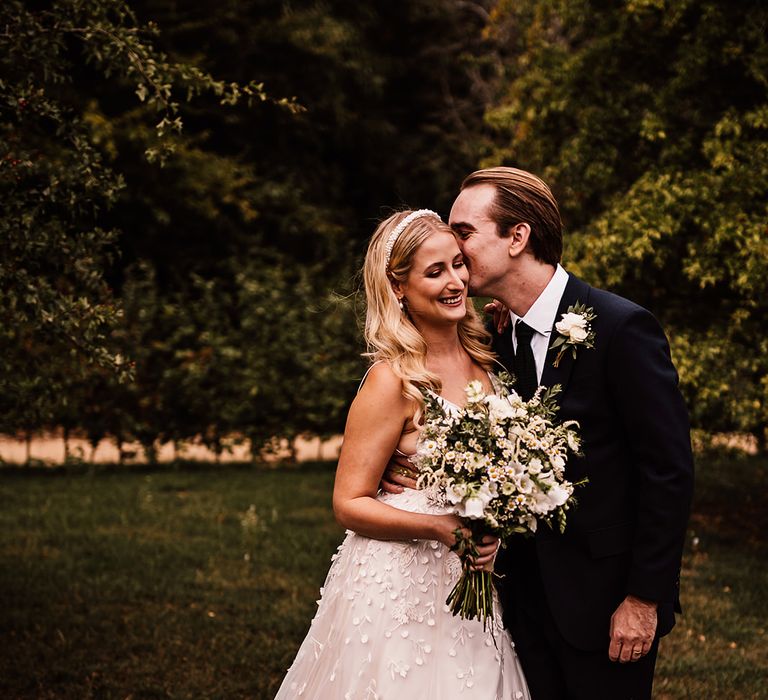 Groom in a navy blue suit kisses the bride in an applique wedding dress with flower design 