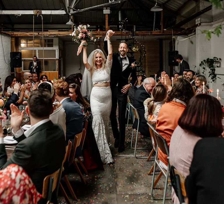 Bride in boho two piece lace bridal outfit and groom in black tux with velvet look bow tie cheering at wedding reception