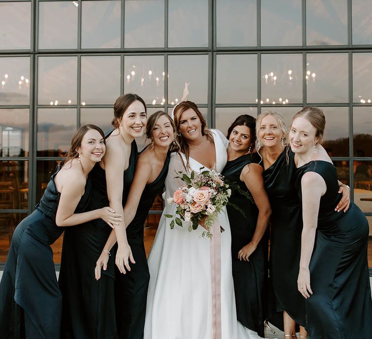 Bride holds pastel bouquet tied with pink ribbon and stands with her bridesmaids in green satin bridesmaid dresses with one shoulder