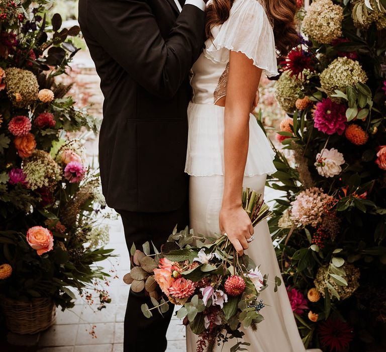 Bride and groom embracing by large luxe floral arch with white and pink carnations, garden roses, red strawflower, eucalyptus, baby’s-breath and various dried flowers 