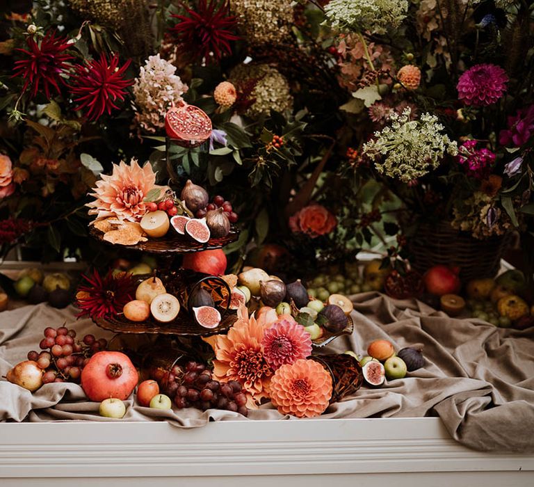 Wedding fruit tower on dark burgundy opaque cake stand surrounded by pink roses, baby's-breath, eucalyptus, carnations, dahlia pinnatas and foliage