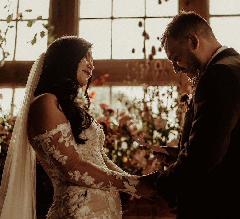 Bride in off the shoulder bodycon wedding dress with long, sheer sleeves and lace detailing holding hands with groom wearing a dark suit with flower patterned tie and pocket square and dried flower boutonniere at the alter of The Giraffe Shed 