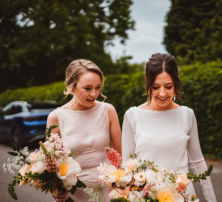 Bride in long satin wedding dress with bridal cover up semi sheer crop top and bridesmaids in light pink satin look bridesmaid dresses holding tropical bouquet with pink garden roses, foliage and baby's-breath 